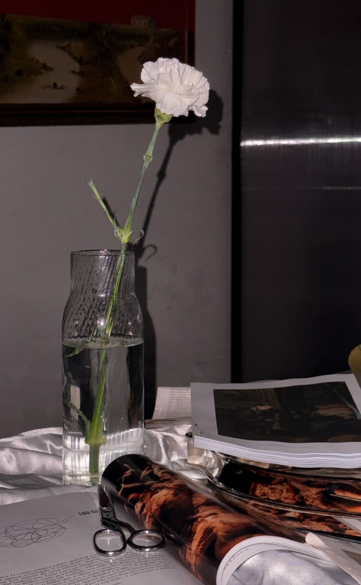 The table at close range under candlelight: a single white carnation in a ribbed glass, a stack of cut magazine pages, scissors, and a dark painting on the wall behind.