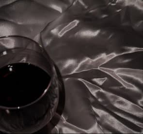 A glass of red wine standing on crumpled silver satin, the fabric catching hard specular highlights.
