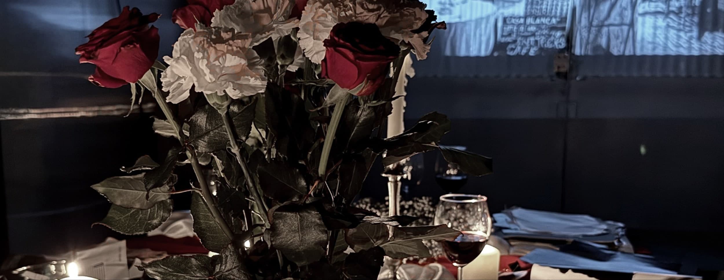 The table seen wide: dark red roses and white flowers massed at the centre, brass candlesticks and wine glasses around them, a black-and-white film projected on the wall behind.